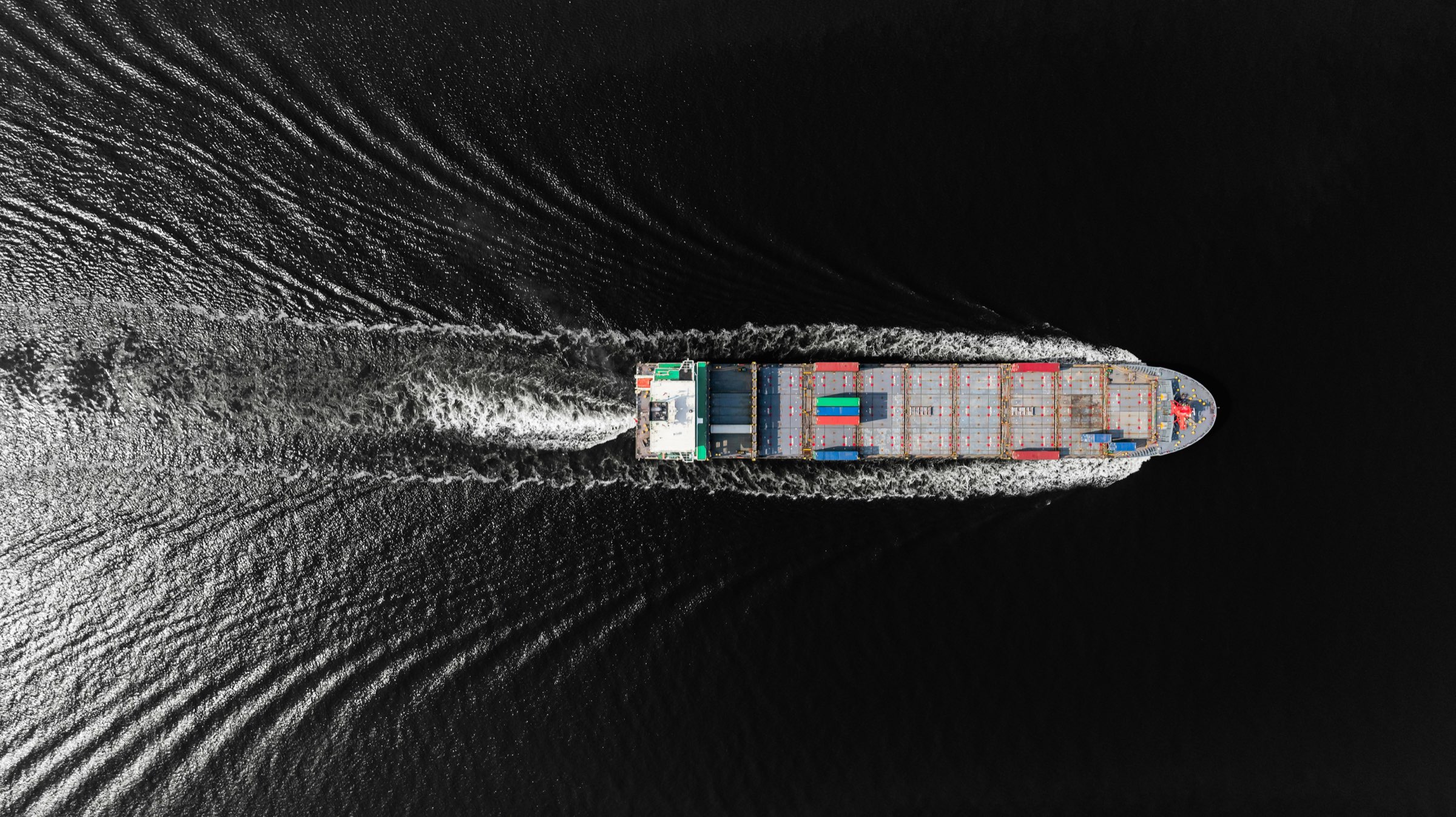 A container ship seen from directly above, its white wake fanning out behind it across near-black water.