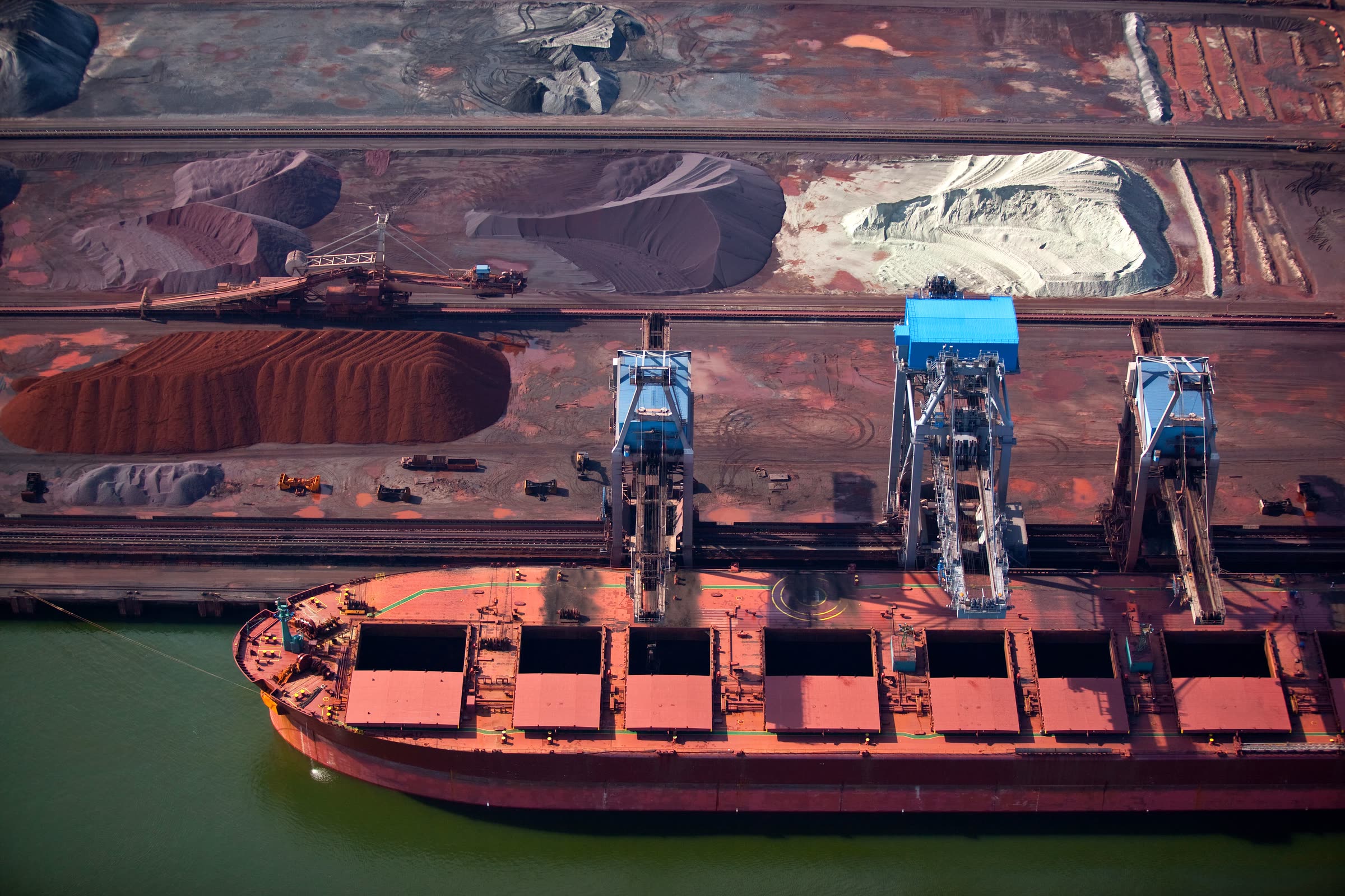 A dry-bulk carrier loading at a terminal, seen from above, with open cargo holds, shiploaders and mineral stockpiles.